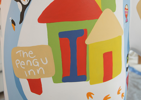 Painted illustration by Gina Baker of the PenguInn a penguing house. Signpost, jumping colourful fish showing the way. Footprints. Penguin swimming backstroke in the water